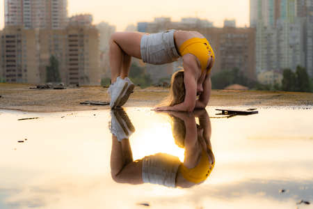 Flexible female gymnast doing handstand and calisthenic with reflection in the water on cityscape background during dramatic sunsetの写真素材