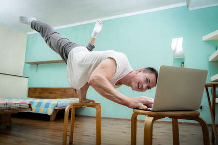 A muscular man keeps balance on one hand and works at a laptop during covid-19. Self-isolation, teleworking and home fitness conceptの写真素材