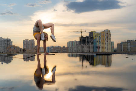 Flexible female gymnast doing handstand and calisthenic with reflection in the water on cityscape background during dramatic sunsetの写真素材