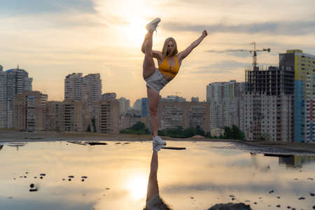 Flexible female gymnast standing split with reflection in the water during dramatic sunset with cityscape backgroundの写真素材