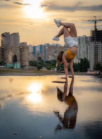 Flexible female gymnast doing handstand and calisthenic with reflection in the water on cityscape background during dramatic sunsetの写真素材