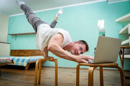 A muscular man keeps balance on one hand and works at a laptop. Self-isolation, teleworking and body training conceptの写真素材