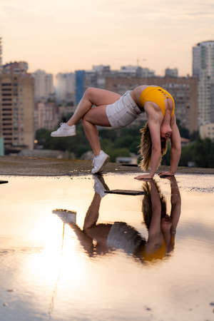 Flexible female gymnast doing handstand and calisthenic with reflection in the water on cityscape background during dramatic sunsetの写真素材