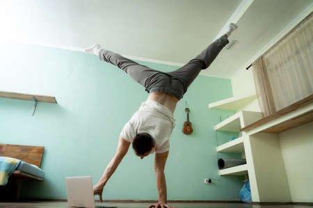A muscular man keeps balance on one hand and works at a laptop. Self-isolation, teleworking and body training conceptの写真素材