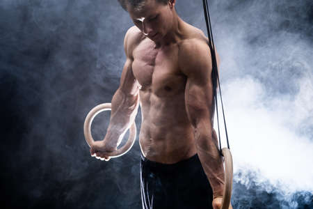 Muscular build man doing calisthenics on gymnastics rings indoor on black, smoked background. concept of motivation, desire and passionの写真素材