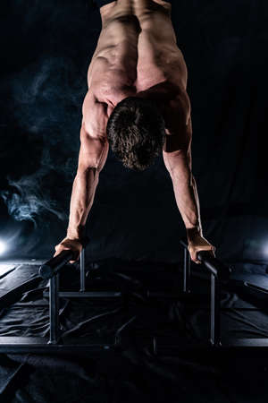 Muscular build man doing crossfit on parallels bar indoor on black, smoked background. concept of healthy lifestyle and calisthenicの写真素材