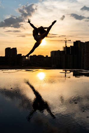 Silhouette of Female flexible dancer jumping during sunset on cityscape background with reflection in the water. Concept of motivation and movementの写真素材