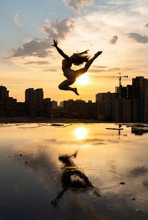 Silhouette of Female flexible dancer jumping during sunset on cityscape background with reflection in the water. Concept of motivation and movementの写真素材