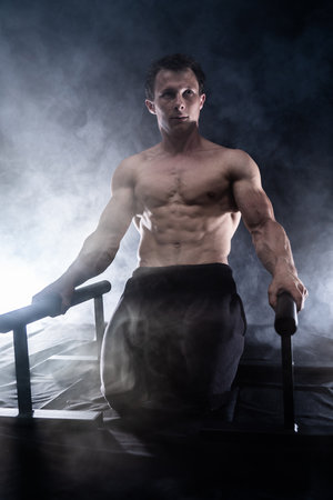 Muscular build man doing calisthenics on parallels bar indoor on black, smoked background. concept of healthy lifestyle and powerの写真素材