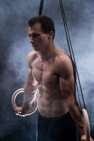 Muscular build man doing calisthenics on gymnastics rings indoor on black, smoked background. concept of healthy lifestyle and power.の写真素材