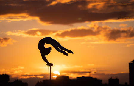 Flexible female circus Artist keep balance and doing contortion on the rooftop against dramatic sunset and cityscape. Handstand and equilibrium conceptの写真素材