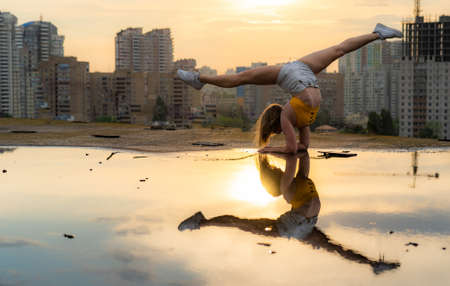 Flexible female gymnast doing handstand and calisthenic with reflection in the water on cityscape background during dramatic sunsetの写真素材