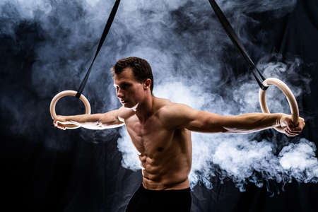 Muscular build man doing calisthenics on gymnastics rings indoor on black, smoked background. concept of motivation, desire and passionの写真素材