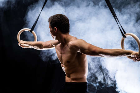 Muscular build man doing calisthenics on gymnastics rings indoor on black, smoked background. concept of healthy lifestyle and power.の写真素材