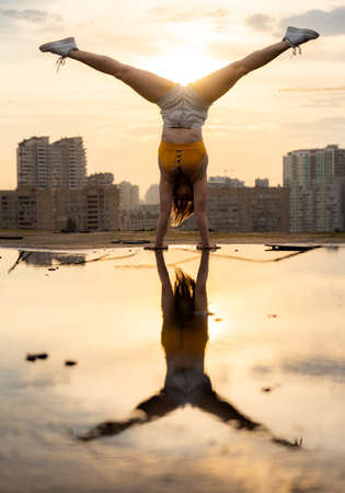 Flexible female gymnast doing handstand and calisthenic with reflection in the water on cityscape background during dramatic sunsetの写真素材