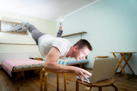 A muscular man practice yoga on one hand and works at a laptop. Self-isolation, remote working and healthy lifestyle conceptの写真素材