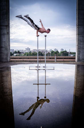 Flexible male circus Artist keep balance by one hand in the concrete structure on sky background with reflection in the water. Equality, consistency and symmetry concept.の写真素材