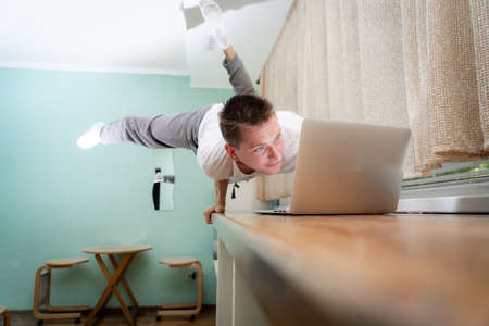 A muscular man keeps balance on one hand and works at a laptop during covid-19. Self-isolation, teleworking and home fitness conceptの写真素材