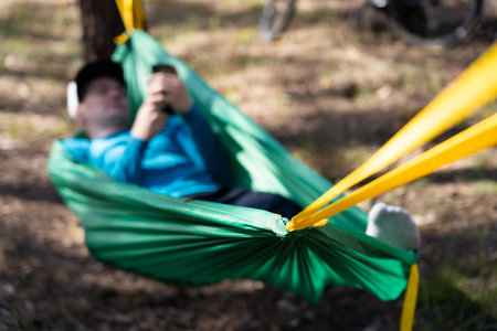 Young happy man using mobile phone in hat relaxing outside in hammock in forest.の写真素材