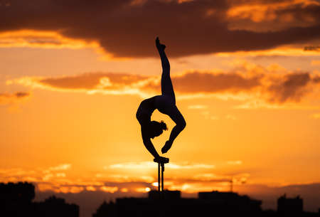 Flexible female circus Artist keep balance and doing contortion on the rooftop against dramatic sunset and cityscape. Handstand and equilibrium conceptの写真素材