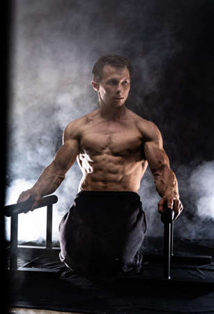 Muscular build man doing crossfit on parallels bar indoor on black, smoked background. concept of healthy lifestyle and calisthenicの写真素材