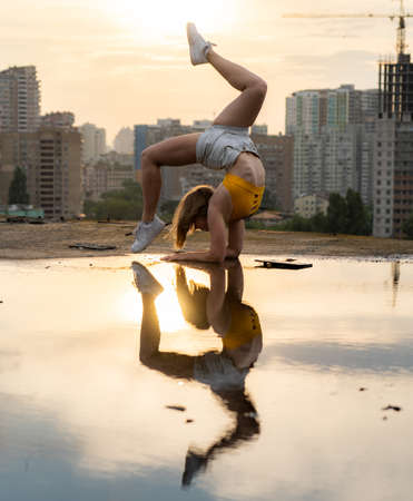Flexible female gymnast doing handstand and calisthenic with reflection in the water on cityscape background during dramatic sunsetの写真素材