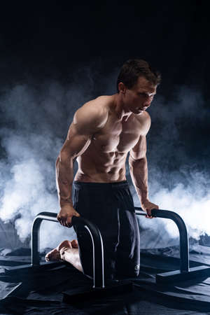 Muscular build man doing calisthenics on parallels bar indoor on black, smoked background. concept of healthy lifestyle and powerの写真素材