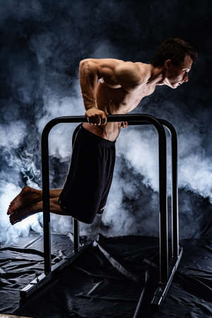 Muscular build man doing calisthenics on parallels bar indoor on black, smoked background. concept of motivation, desire and passionの写真素材