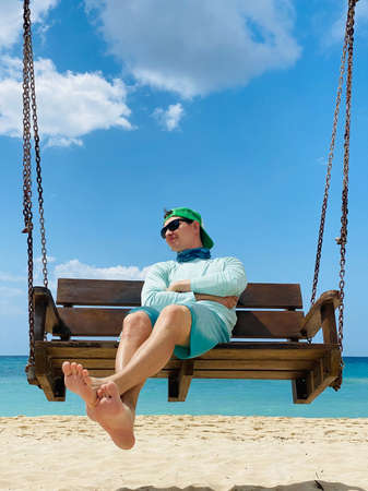 Travel lifestyle in summer season. Young Asian man photographer sitting on wooden swing at tropical beach. Relaxation and carefree concepts.の写真素材