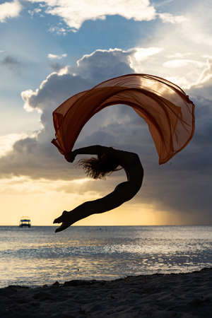 Flexible fit woman jumping with silk during dramatic sunset with stormy clouds. Concept of individuality, creativity and self-confidenceの写真素材