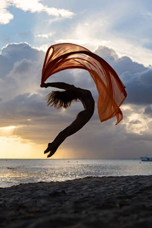 Silhouette of flexible fit woman jumping with silk during dramatic sunset with stormy clouds on the seascape background. Concept of happiness, freedom and carefree.の写真素材