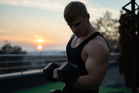 Caucasian white muscular man lifting dumbbells against the sunset sky background. Concept of willpower, motivation and healthy lifestyleの写真素材