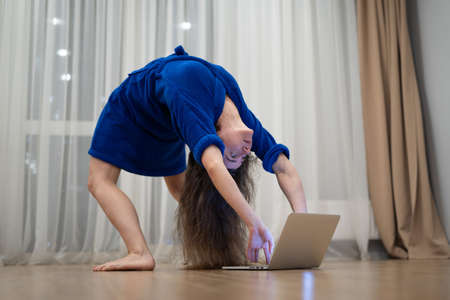 Very flexible woman exercising at home in front of her laptop, stretching her back. Concept of individuality, creativity and self-confidenceの写真素材