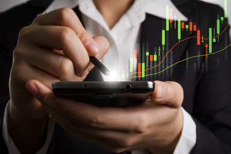 Close up business woman hand holding smart phone and pen with candle graph for trade stock market .の写真素材