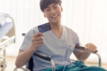 Young teenage Asian man holding credit card sitting on the bed in the hospital with smiley face.の写真素材