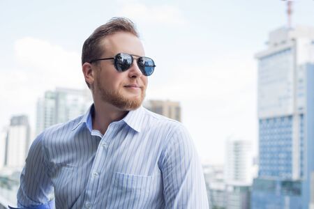 Young caucasian man wearing sunglasses being outdoor at the roof top of high building looking at the city view.の写真素材
