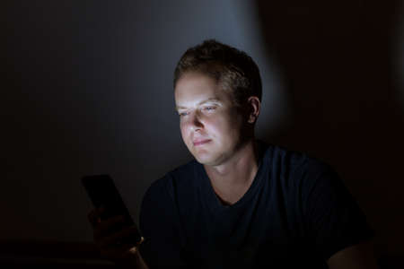 Young attractive Caucasian man using smart phone in the dark room, risky for his eye's health.