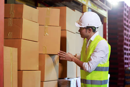 Young attractive Asian engineer worker wearing white hard hat and safety vest looking at the pile of cardboard boxes at the warehouse work place.の写真素材