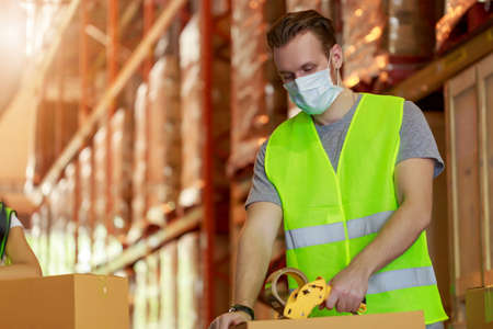 Young caucasian employee male packing cardboard box with scotch tape, wearing protective face mask, new normal after pandemic crisis.の写真素材
