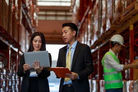 Asian Warehouse manager and female worker interacting talking checking inventory with engineer worker standing behind, manager team working at the storehouse.の写真素材