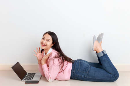 Young attractive collage student lying down on the floor by the wall learning online at home, Asian...
