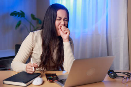 Young sleepy Asian woman sitting in front of computer yawning over worked at night, Asian female...