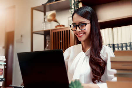 Beautiful happy Asian woman sitting in front her computer laptop at the wooden working table over...