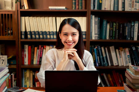 Beautiful happy Asian woman sitting in front her computer laptop at the wooden working table over...