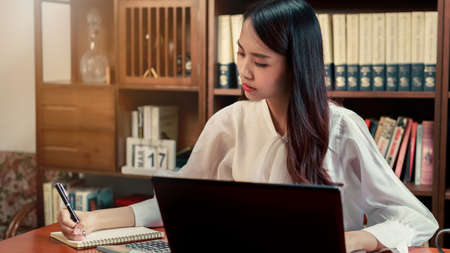 Beautiful Asian woman writing the notes while  sitting in front her computer laptop at the wooden...