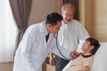 Asian Pediatrician man doctor consulting sick girl patient listening lungs using medical stethoscope during disease examination in hospital ward. Hospitalized little child recovering after surgery.の写真素材