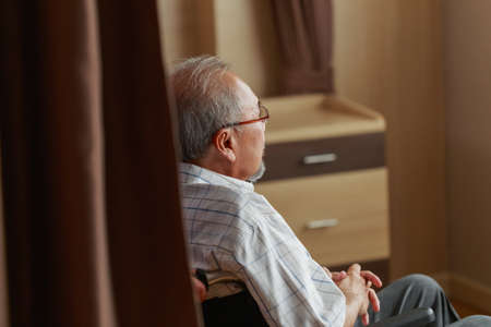 Rear view of senior Asian male patient looking outside the window while sitting in wheelchair in bedroom at retirement home, Social distancing and self isolation.の写真素材
