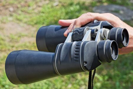 hand holds big black binoculars on the street against the background of green grass