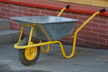 gray yellow metal wheelbarrow stands on the pavement near a brick wallの写真素材
