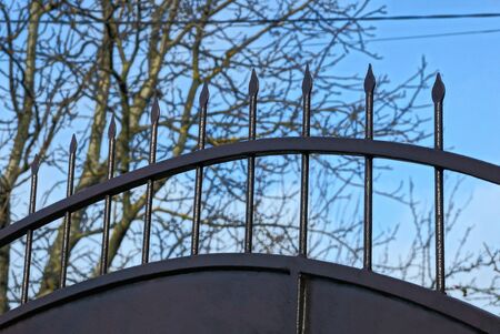 part of a black iron fence with sharp rods against the blue sky and gray branches